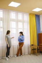 Two women are talking in a bright room featuring large windows with flowing curtains in yellow and