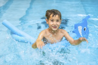 Smiling boy having fun in a swimming pool, playing with a pool noodle float shaped like a shark,