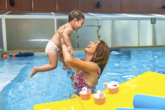 Happy mother lifting her laughing baby out of the water in an indoor swimming pool next to floating