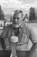 Elderly gentleman with beard and sunglasses in a garden restaurant with a drink, black and white,