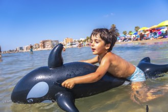 Happy child riding on an inflatable orca toy in the sea, splashing through waves and enjoying a