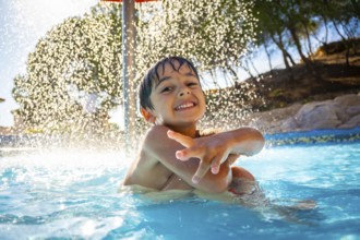 Smiling boy enjoying playful moments under a refreshing water shower in a swimming pool on a