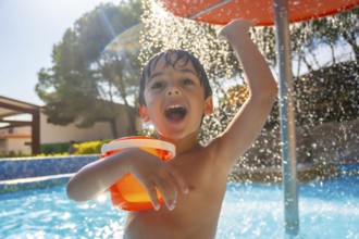 Boy holding orange bucket and raising arm under refreshing water shower in swimming pool on sunny
