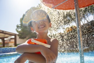 Smiling child holding a bucket and enjoying refreshing shower in a swimming pool on a sunny summer