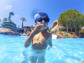 Young boy wearing sunglasses blowing a kiss while standing in a swimming pool at a water park with