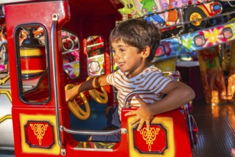 Young boy smiling while steering a vibrant red amusement park ride, surrounded by colorful