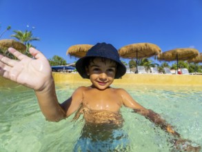 Smiling child in a sun hat waves while playing in a clear swimming pool, surrounded by straw