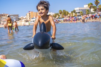 Smiling boy riding an inflatable orca toy in the sea waves, enjoying summer vacation at a crowded
