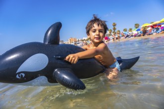 Young boy enjoying summer vacation, riding an inflatable orca toy in the ocean at a crowded beach