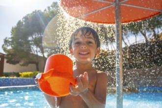 Smiling child enjoying a sunny day at the pool, holding an orange bucket under a refreshing water