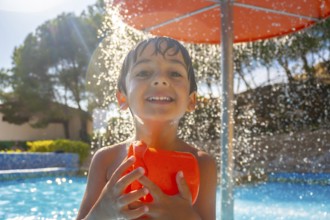 Smiling boy holding an orange water toy under a splash of water, enjoying a sunny day by the pool
