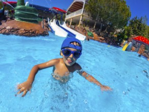 Young boy wearing sunglasses and a blue cap, enjoying a summer day at a water park, swimming in the