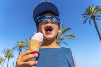 Young boy with sunglasses and cap enjoying a strawberry ice cream cone on a hot summer day with