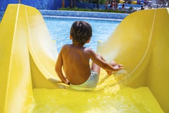 Young boy enjoying summer holidays sliding down yellow waterslide into swimming pool in aqua park