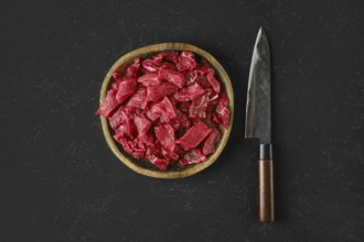A wooden bowl holds freshly cut pieces of raw beef, showcasing a vibrant red color. Beside the