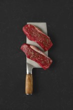 Two raw beef steaks seasoned with spices on a large metal cleaver with a wooden handle. The dark