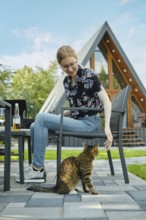 A girl plays with a cat while sitting at a picnic table on the lawn of a country house