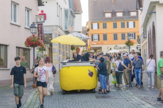 People stroll through a narrow alley in the old town centre, flanked by shops and flowers, 950th