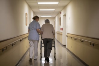 Back view of nurse helping older woman walk with walker in rehabilitation center hallway. Elder