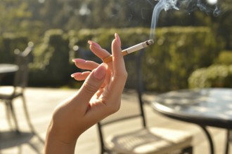 Elegant female hand with long pink nails holding a burning cigarette. Summer sunlight in outdoor