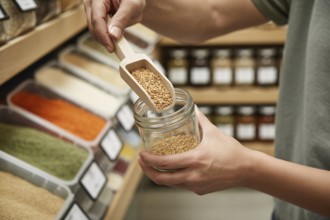 Hands filling small glass jar with grains using wooden scoop at bulk store. Concept of zero-waste