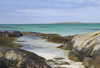 Rocky coastline with turquoise sea and gentle waves under a blue sky, Eriksay, South Uist,