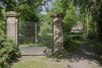 A wrought-iron gate between two brick pillars in a green, shady landscape with a building in the
