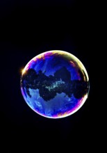 A shimmering and floating soap bubble in the colours of the rainbow against a black background,