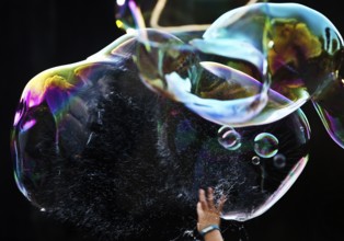 A hand bursts a large iridescent soap bubble against a black background, Recklinghausen, Germany