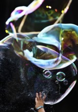 A hand bursts a large iridescent soap bubble against a black background, Recklinghausen, Germany