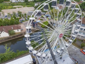 Aerial view of a Ferris wheel next to a river with cars on the road and trees in the background,