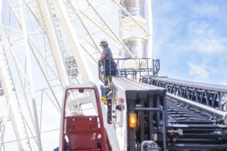 Firefighter on a lifting platform next to a Ferris wheel in front of a clear sky, fire brigade