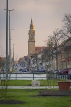 City scene with church tower in the background, surrounded by cars and buildings along a street at
