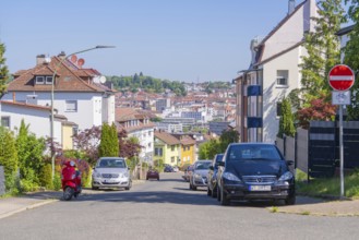 Urban street with cars, a no cycling sign, a scooter and a backdrop of houses under a clear sky,