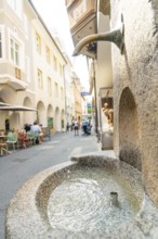 A stone fountain with running water in an alley next to a café, Merano, South Tyrol, Italy
