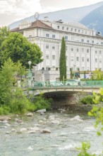 Historic riverside hotel with bridge and lush vegetation against a mountain backdrop, Merano, South