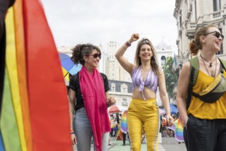 Three women walking and smiling during a vibrant gay pride parade, one raising her fist in a