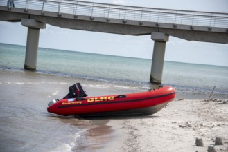A DLRG lifeboat lies on the beach in front of a bridge with walkers and the sea in the background,
