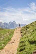 A hiker on a mountain path with outstretched arms in front of an impressive mountain scenery,
