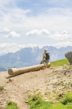 A hiker sits on a tree trunk and looks at the surrounding mountains, Schlehrn hike, Alpe di Siusi,