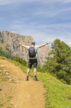 A man stands joyfully with outstretched arms on a sunny mountain path, Schlehrn hike, Alpe di