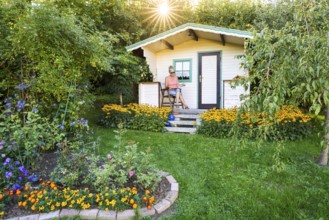 A woman sits relaxed on the small veranda of a garden shed and reads. Blooming summer flowers in