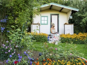 A woman in a dress sits relaxed on the small veranda of a garden shed and reads. Blooming summer