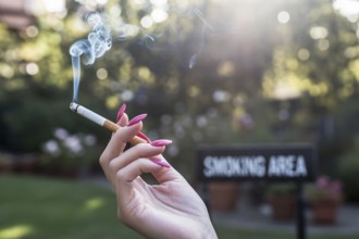 Woman's hand with long pink nails holding a cigarette. Blurred smoking area sign in background.