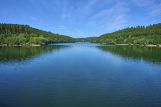 A wide view over a calm lake surrounded by dense forests under a blue sky, Oleftalsperre,