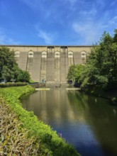 Dam wall with river in the foreground, surrounded by trees and reflected in the water,