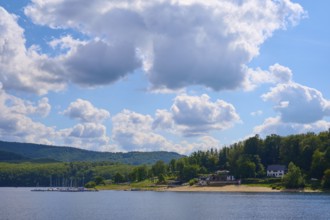 Houses and boats on the shore of a lake, surrounded by green hills under a cloudy sky, summer,