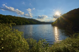 Sunbeams reflecting in the lake surrounded by wooded hills under a blue sky with clouds, summer,