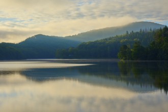 Calm lake with mountains in the background, dense forest and morning sun, gentle mist on the shore,