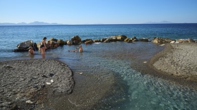 People swimming in the calm blue sea by rocks and sandy shore, Embros Thermal Baths, Kos,